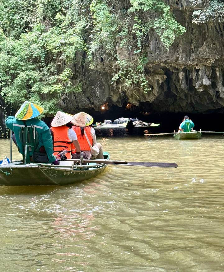 Tourists in conical hats paddle a small boat toward a dark limestone cave entrance on a calm river.