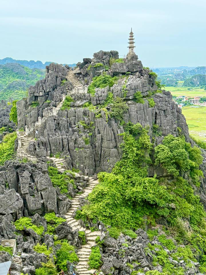 Steep rocky peak covered with greenery and zig-zag stone steps overlooking rice fields below.