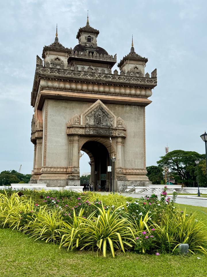 Visitors walk under the ornate arch of Patuxai monument surrounded by gardens.