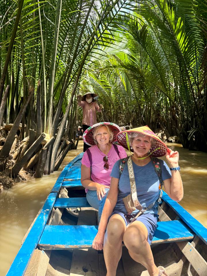 Smiling tourists ride a small wooden boat through dense nipa palms on muddy river water.
