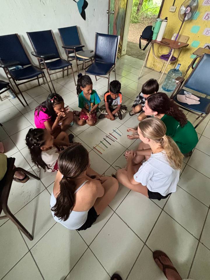 Volunteers and local children sit in a circle on tiled floor playing an educational stick game.