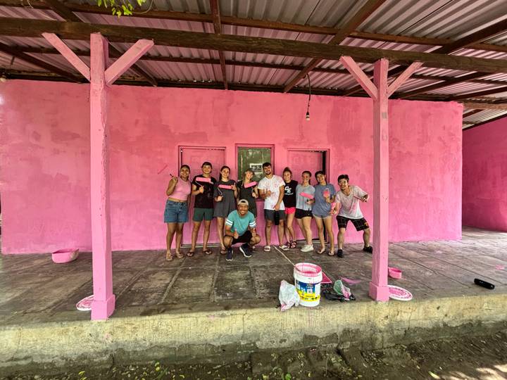 Group of volunteers in paint-splattered clothes pose proudly outside a newly pink-painted building.