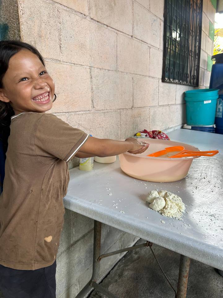 Smiling child kneads dough beside a mixing bowl on an outdoor table.