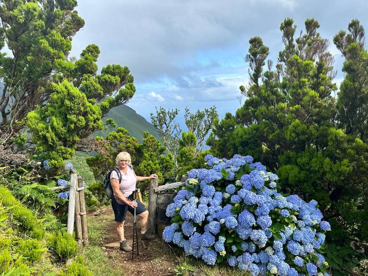 Smiling hiker standing beside blooming blue hydrangeas on a lush mountain trail with distant ocean views under partly cloudy skies.