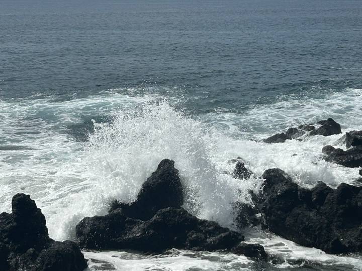 Powerful ocean waves crash dramatically against jagged black volcanic rocks sending white spray into the air.