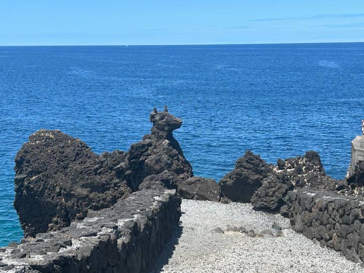 Clear blue Atlantic waters border rugged dark lava rock formations under a bright summer sky.