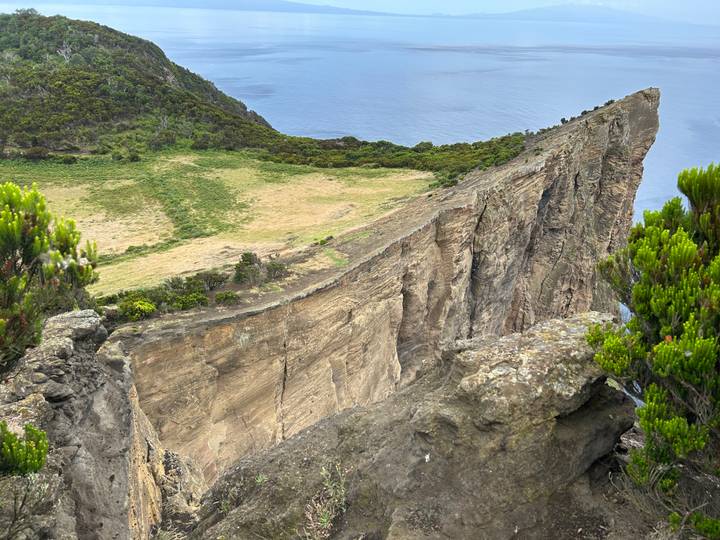 Dramatic sheer cliff edge dropping toward the sea with contrasting green plateau and deep blue water beyond.