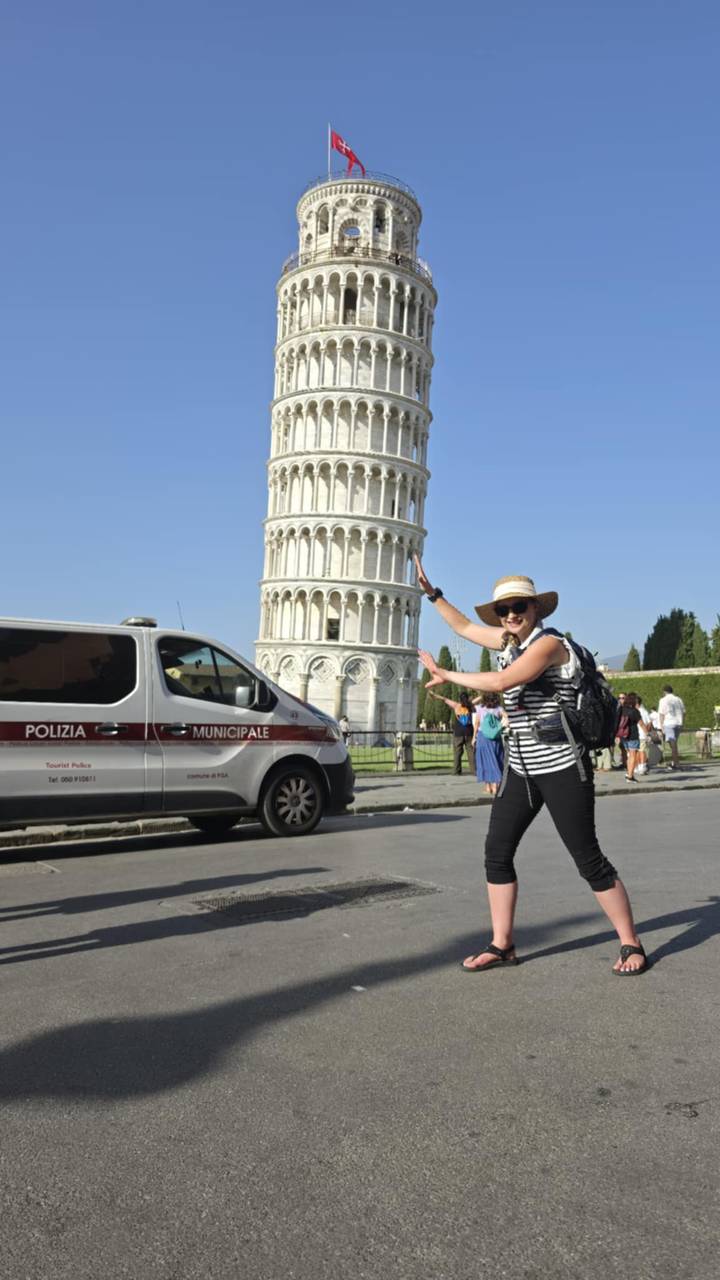 Tourist playfully posing as if supporting the Leaning Tower of Pisa with police van and visitors in the square