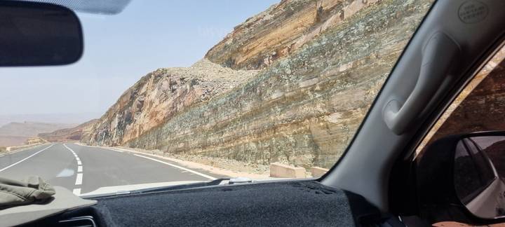 Mountain road seen through a car windshield alongside multicolored rock strata.