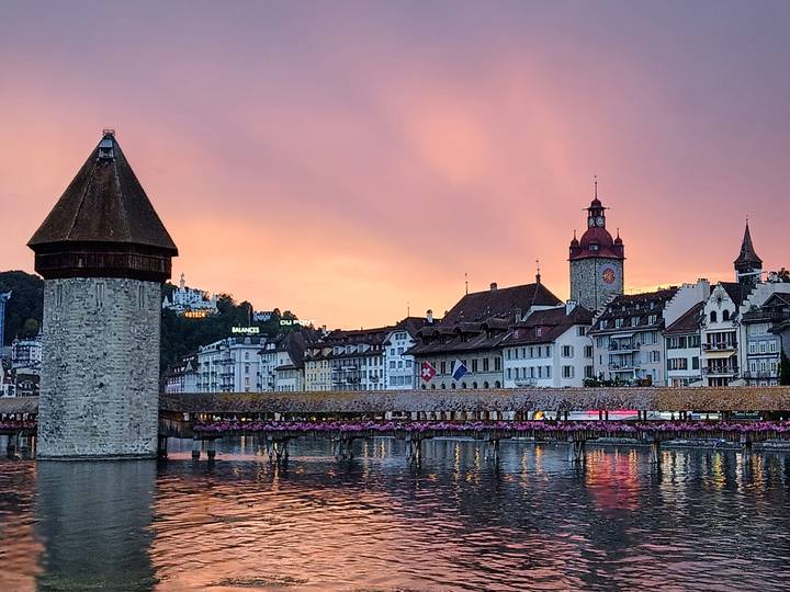 Chapel Bridge and Water Tower reflect in Lake Lucerne beneath a pink sunset sky.