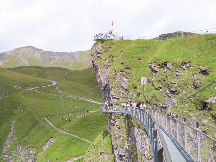 Crowds walk along a dramatic suspended walkway clinging to a grassy cliff face.