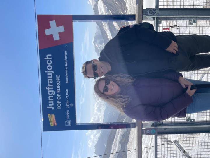 Smiling couple pose beside Jungfraujoch ‘Top of Europe’ sign with snowy alpine peaks behind.