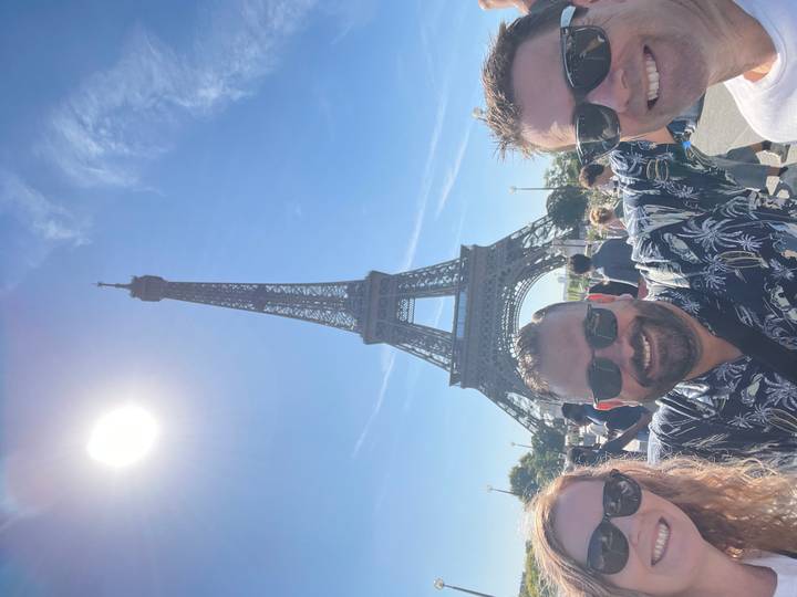 Friends pose with arms wide in front of the Eiffel Tower under a bright midday sun.
