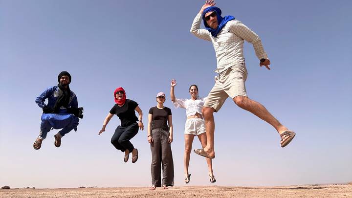 Five travelers in desert scarves leap joyfully against a clear blue sky on sandy terrain.