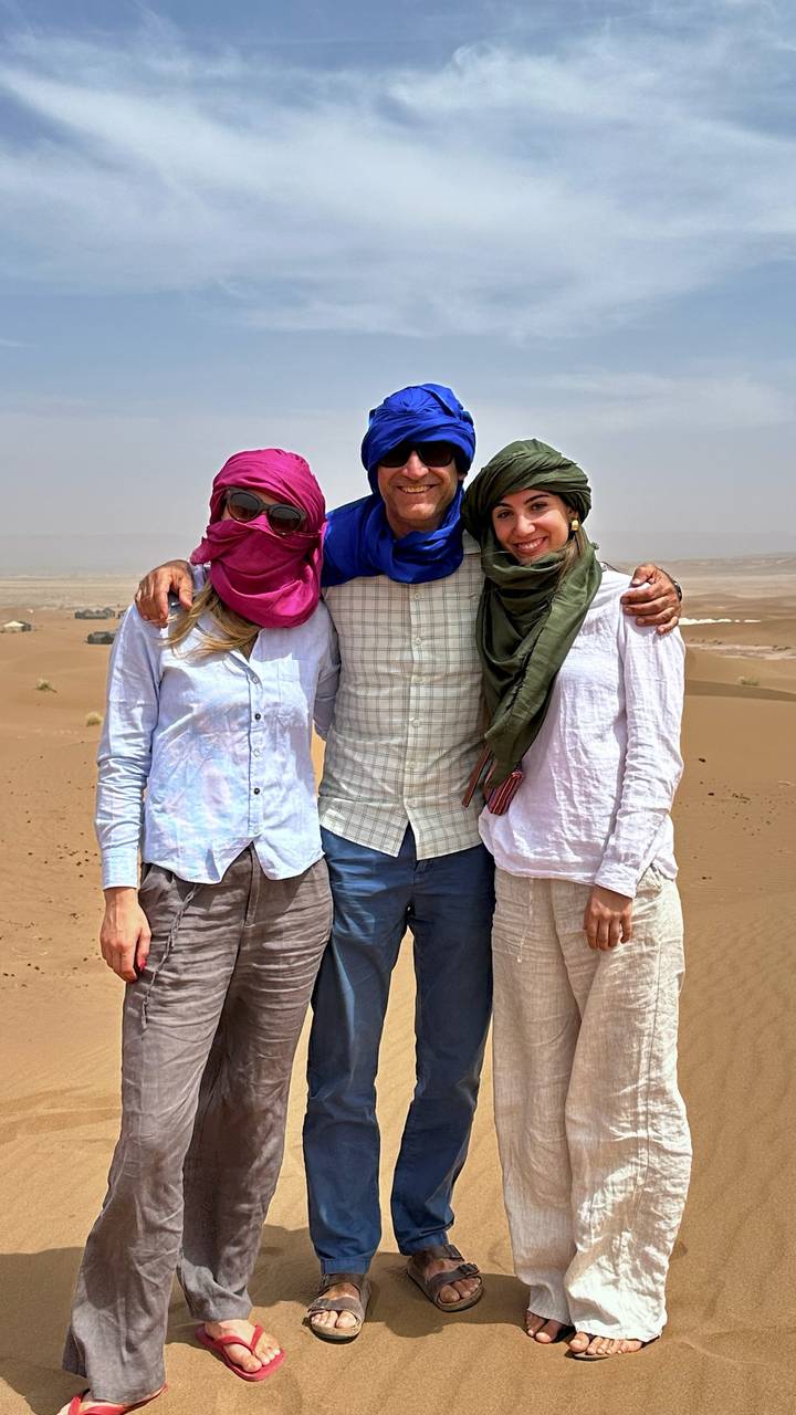 Three travelers wearing headscarves pose arm in arm on a sandy ridge overlooking desert tents.