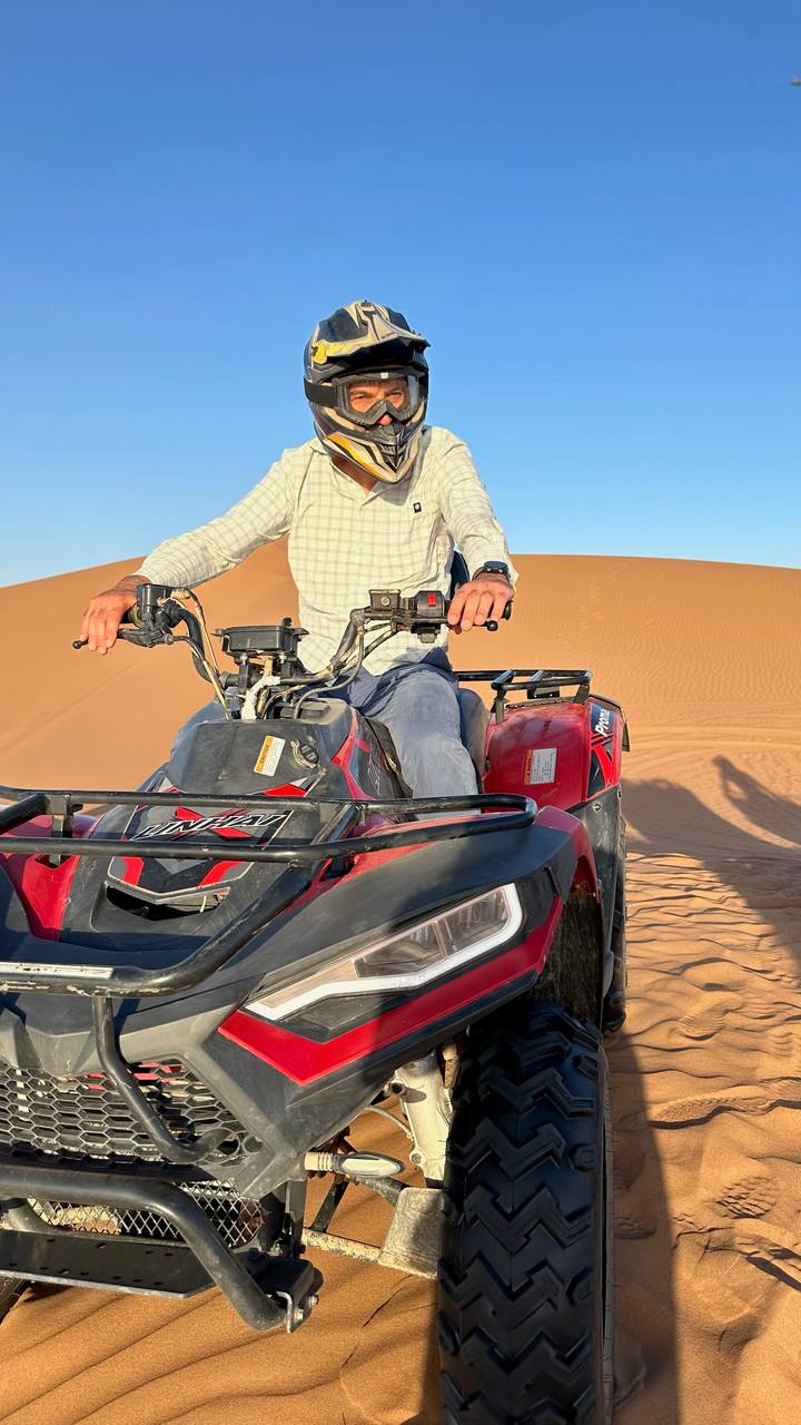 Man drives a red quad bike over smooth orange sand dunes under bright sun.