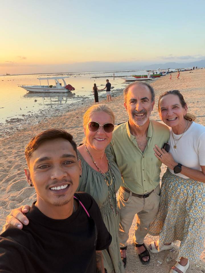 Group of four friends posing on a sandy beach at sunset with a small boat and gentle waves behind.