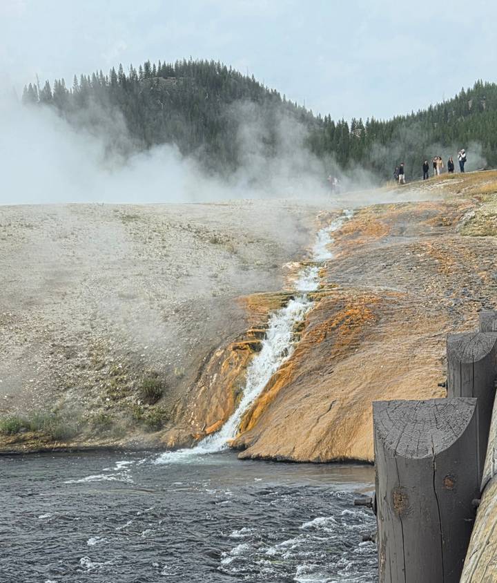 Steaming geothermal run-off flowing over vivid orange mineral deposits in a Yellowstone thermal area with visitors in the distance.