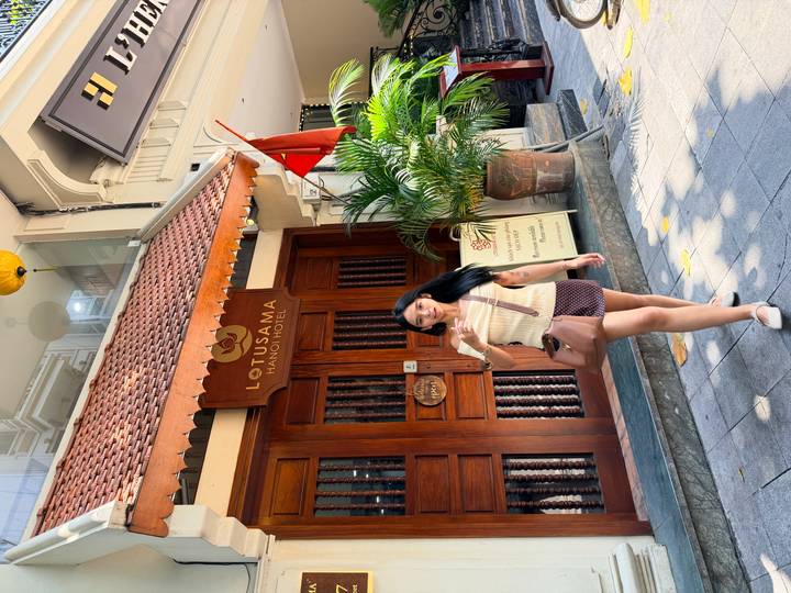 A woman poses in front of the Lotusama Hanoi Hotel entrance along a leafy sidewalk.