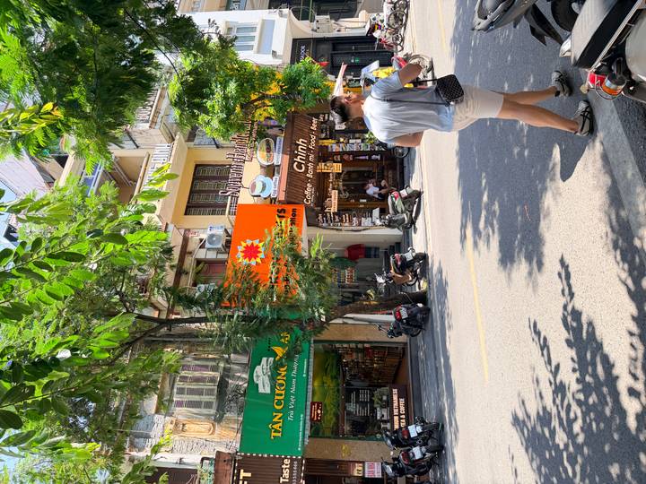 Tree-lined Hanoi street with scooters, shops, and pedestrians under bright midday light.