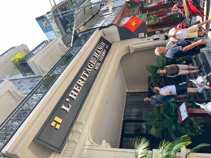 Tourists stand on the steps of L’Heritage Hanoi hotel beneath its black and gold sign.