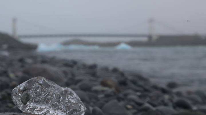 Out-of-focus ice fragment on pebbled shore with bridge and Jokulsarlon lagoon behind in fog.