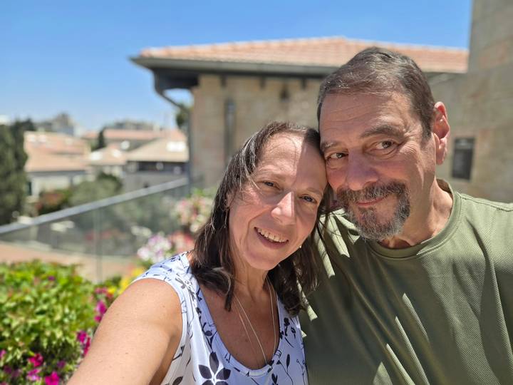 Close-up selfie of a smiling couple with stone buildings and bright flowers softly blurred in the background.