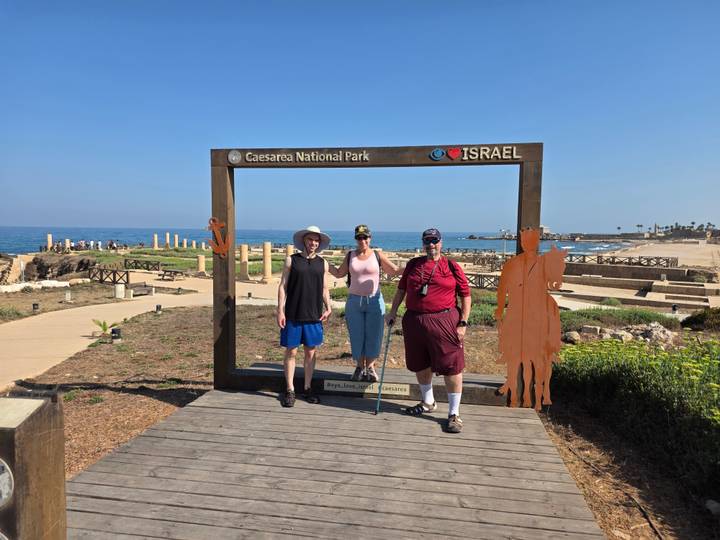 Three travellers pose inside a large wooden picture frame that reads 'Caesarea National Park' with Mediterranean ruins behind.