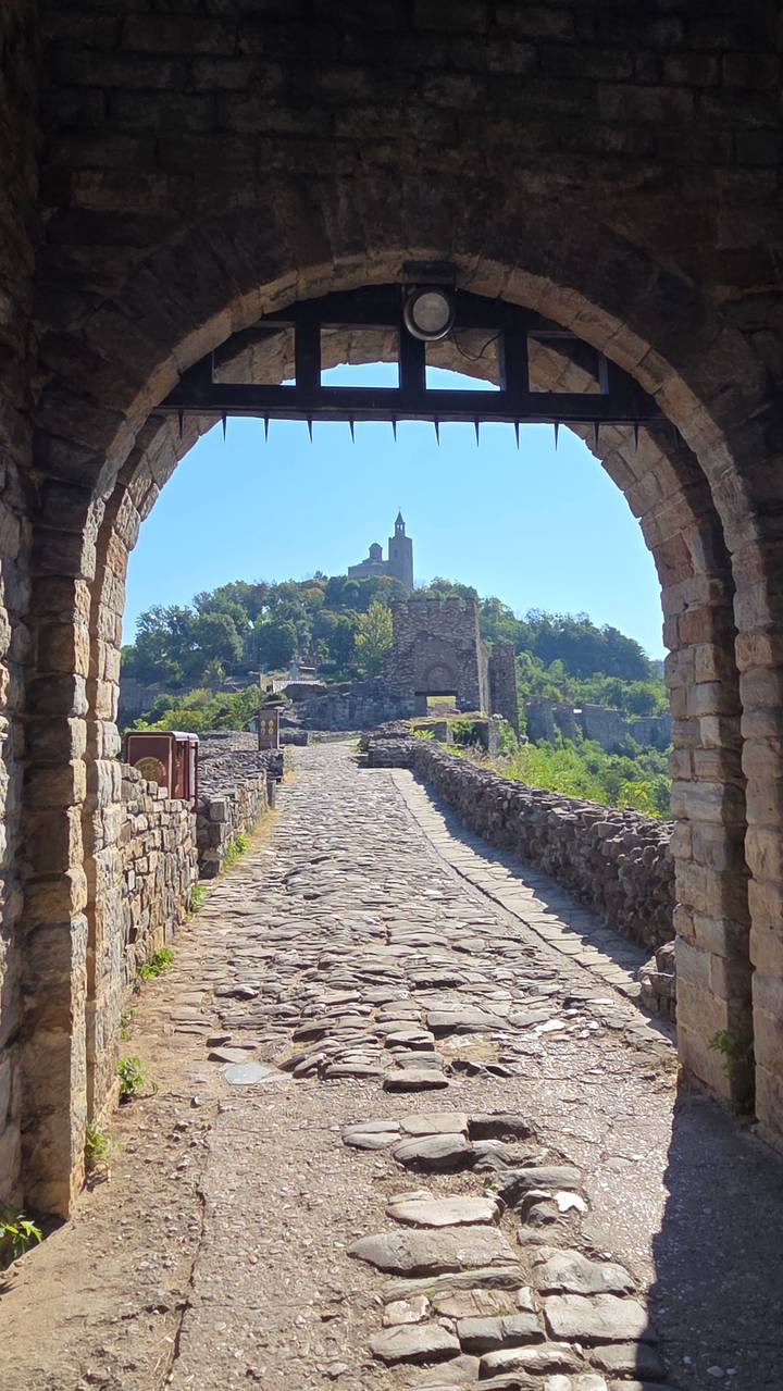 Stone gateway arch framing a cobbled path leading up to the hilltop Tsarevets Fortress tower amid green trees.