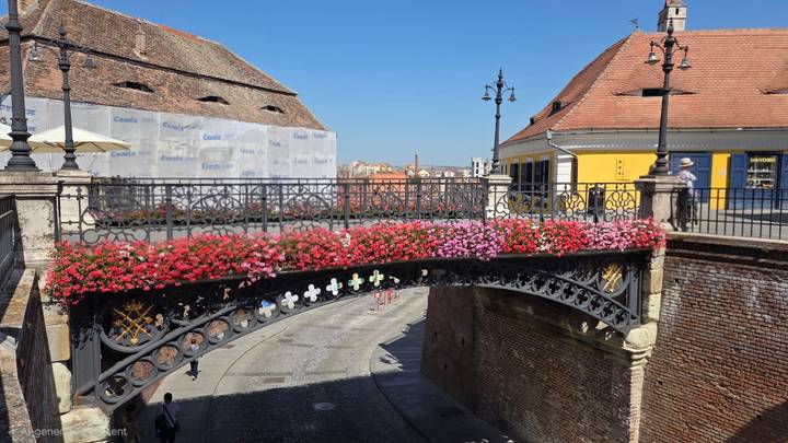 Cast-iron Bridge of Lies decorated with pink and red flowers spanning a cobbled lane between colourful roofs.