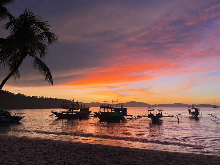 Vibrant sunset with dramatic clouds silhouetting palm trees and several anchored outrigger boats on a serene shoreline.