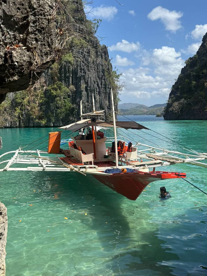 Bright red outrigger boat moored in a turquoise lagoon surrounded by steep limestone cliffs and jungle.