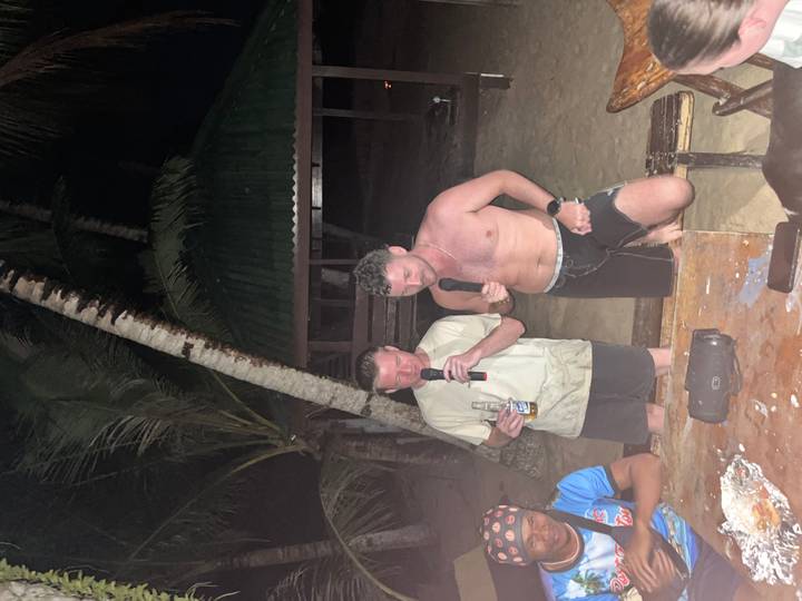 Two men singing karaoke with microphones on a tropical beach at night, illuminated by camera flash.