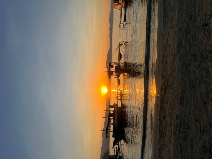 Golden sunrise over calm waters with silhouetted wooden boats and distant hills seen from a sandy beach.
