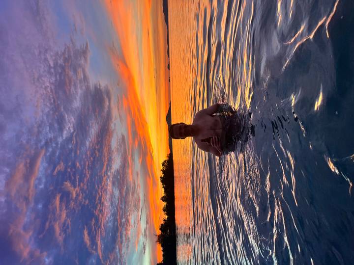 Man waist-deep in shimmering sea at sunset holding a drink, with dramatic orange and purple sky behind.
