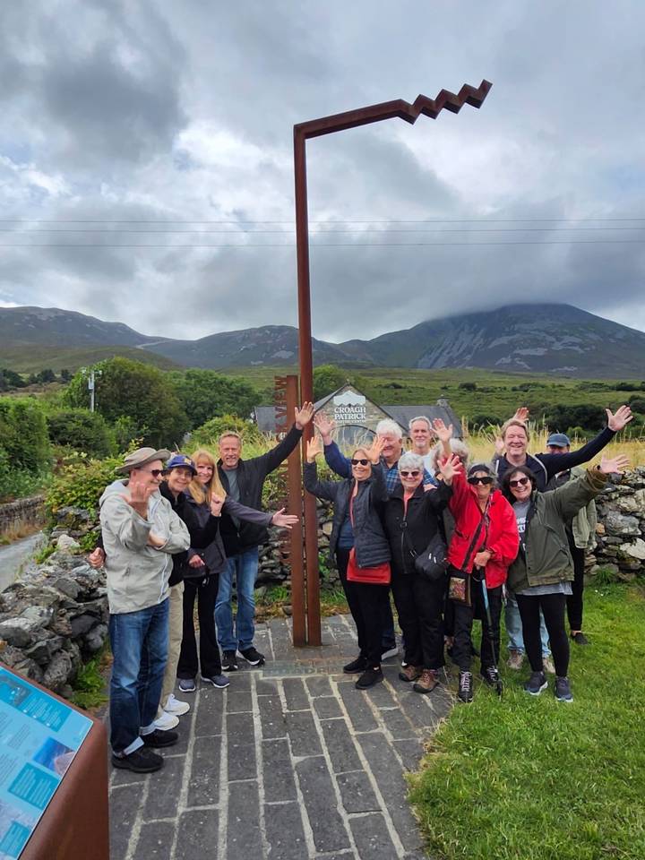 Smiling tour group waving enthusiastically in front of misty green mountains and stone walls in rural Ireland.