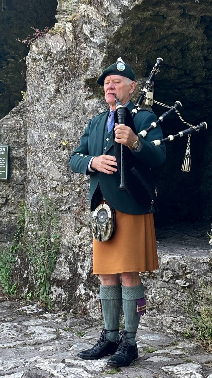 Traditional piper in kilt and jacket playing bagpipes beside ancient stone walls.