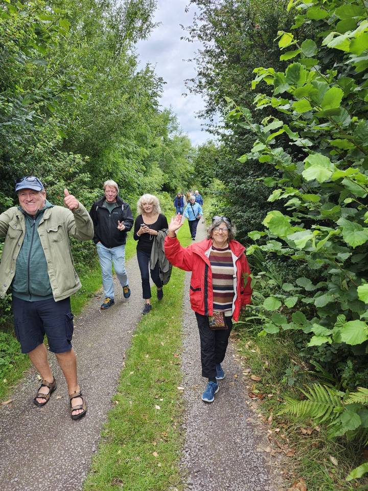 Travellers hiking along a lush green woodland path, smiling and waving.