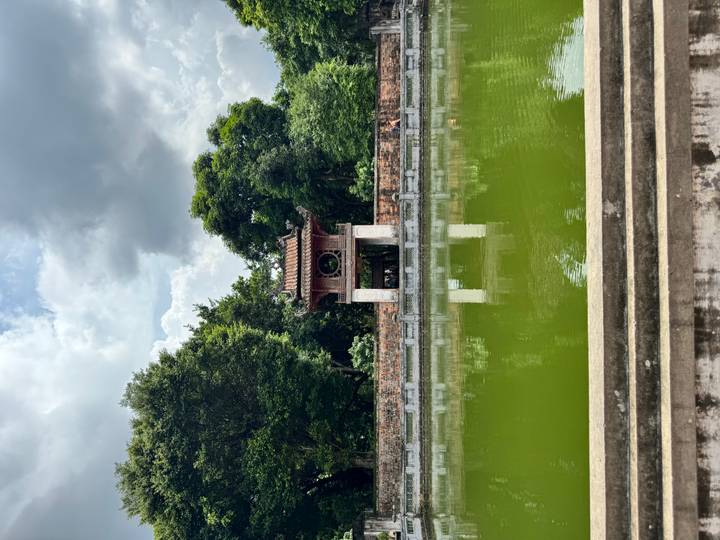 Historic pavilion reflected in a green pond, framed by lush trees and cloudy sky.