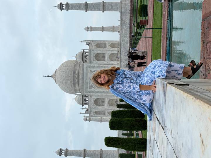 Woman in floral attire poses at marble bench with Taj Mahal rising majestically behind.