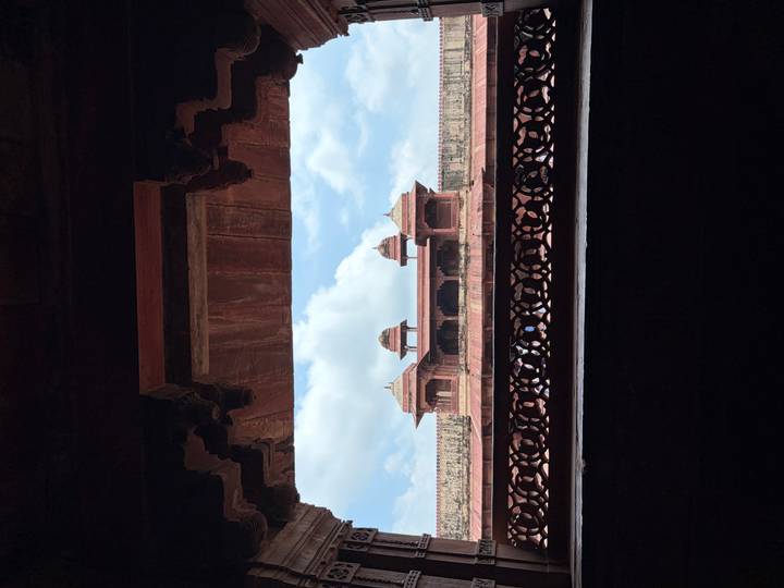 View of a red-sandstone pavilion framed by an ornate arch with blue sky and clouds in the background