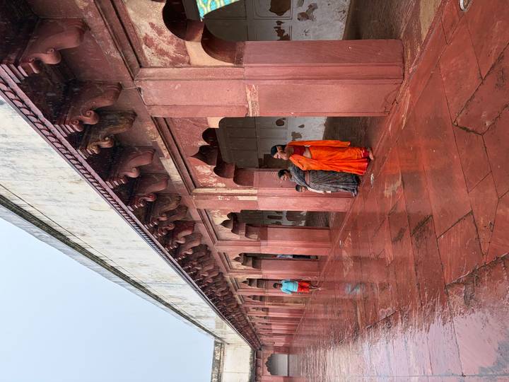 Visitors in colorful saris shelter under red-sandstone arches of a historical corridor on a rainy day