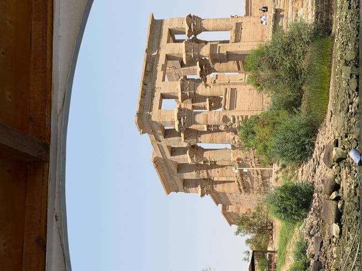 Ancient riverside temple columns of Philae seen from a boat on the Nile under a clear sky.