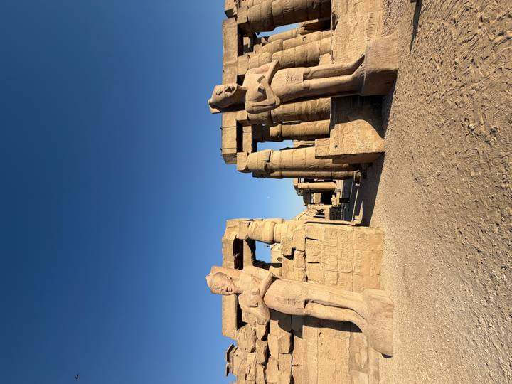 Two colossal pharaonic statues flank an entrance lined with massive pillars under a deep blue sky.