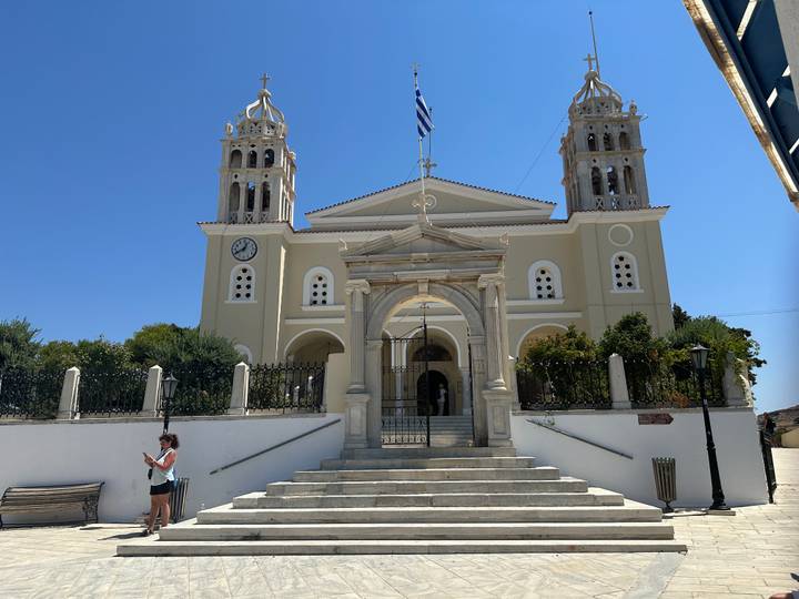 Neo-Classical Greek church with twin bell towers and Greek flag flying overhead.