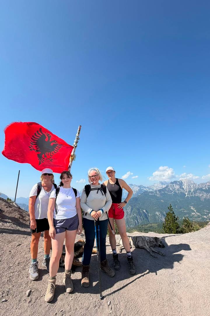 Four hikers posing at a mountain summit beside a large Albanian flag with alpine peaks behind.