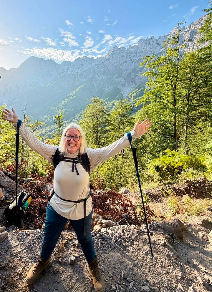 Cheerful hiker with trekking poles raises arms in a lush alpine forest with mountains beyond.