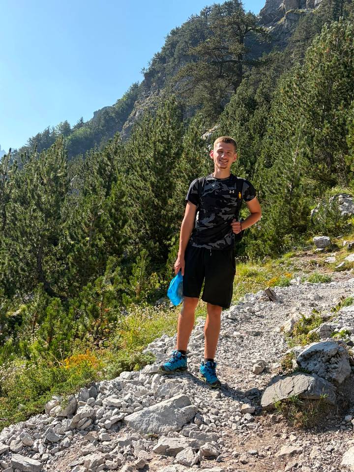 Young man standing on a stony mountain trail surrounded by dense pine trees in Albania.