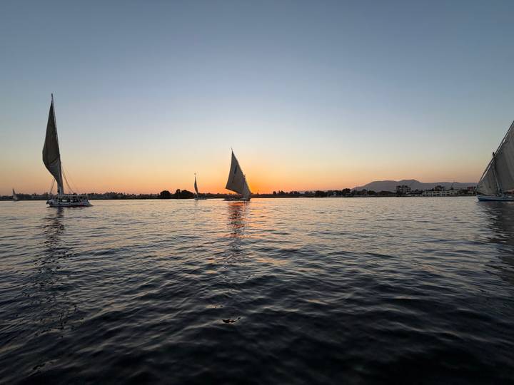 Feluccas sail on the Nile at a vivid orange sunset with rippling reflections on the water.