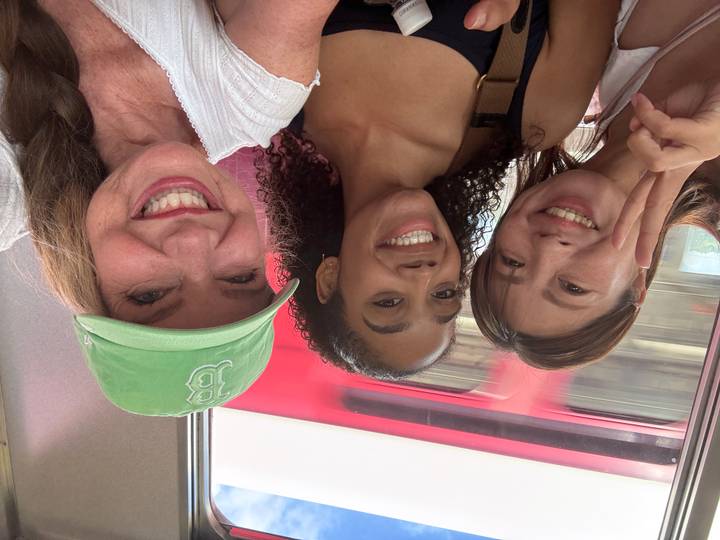 Three women smiling for a selfie inside a moving train in Japan, with a red train blurred outside the window.